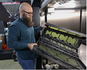 Bald man with a beard wearing glasses and a blue plaid shirt holds a black tray with hexagonal green inserts.