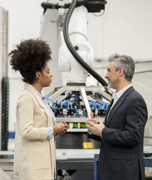 Two professionals in business attire stand in front of a large robotic arm and industrial machinery, engaged in conversation.