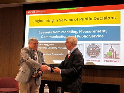 Two men in suits shake hands in front of a projector screen displaying "NC STATE UNIVERSITY" and "Engineering in Service of Public Decisions."