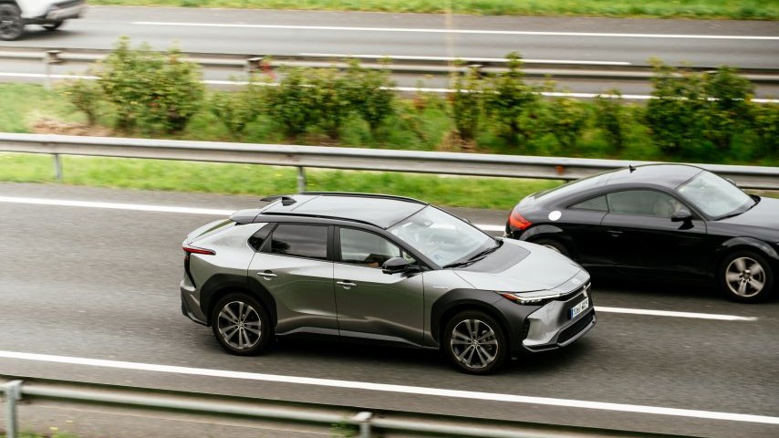 Silver electric SUV driving on a highway alongside other vehicles, with blurred greenery and a guardrail visible.